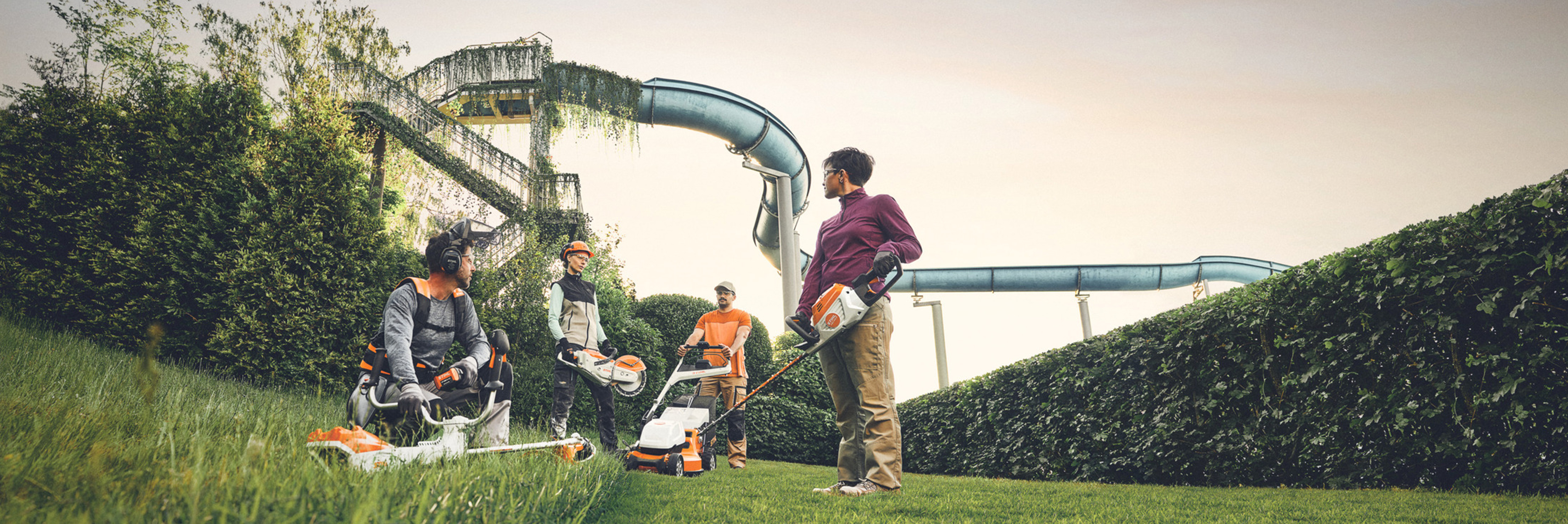 group of people work with STIHL gardening tools in a green area with a large hedge and water slides in the background. They are equipped with brush cutters, lawn mowers and protective equipment.